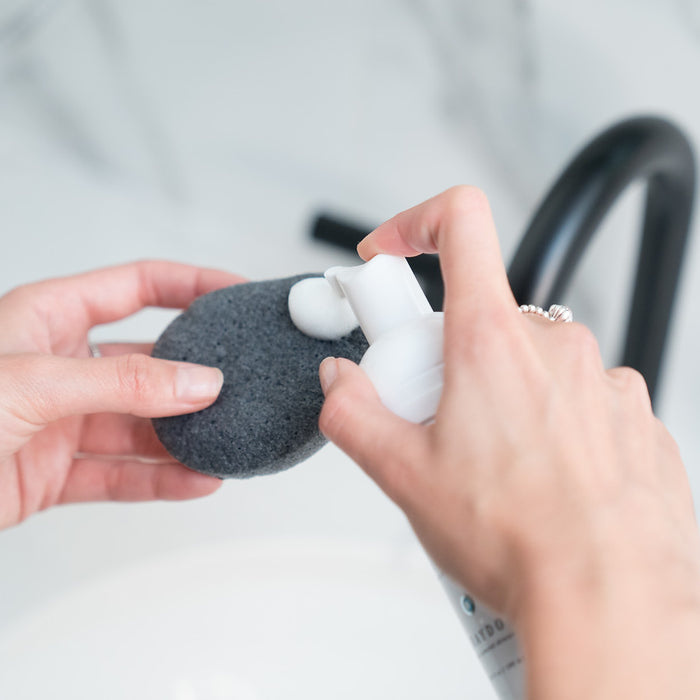 Close-up of a woman dispensing a white foaming cleanser for combination skin onto a black-coloured bamboo charcoal sponge for sensitive skin with a modern-looking sink in the background.