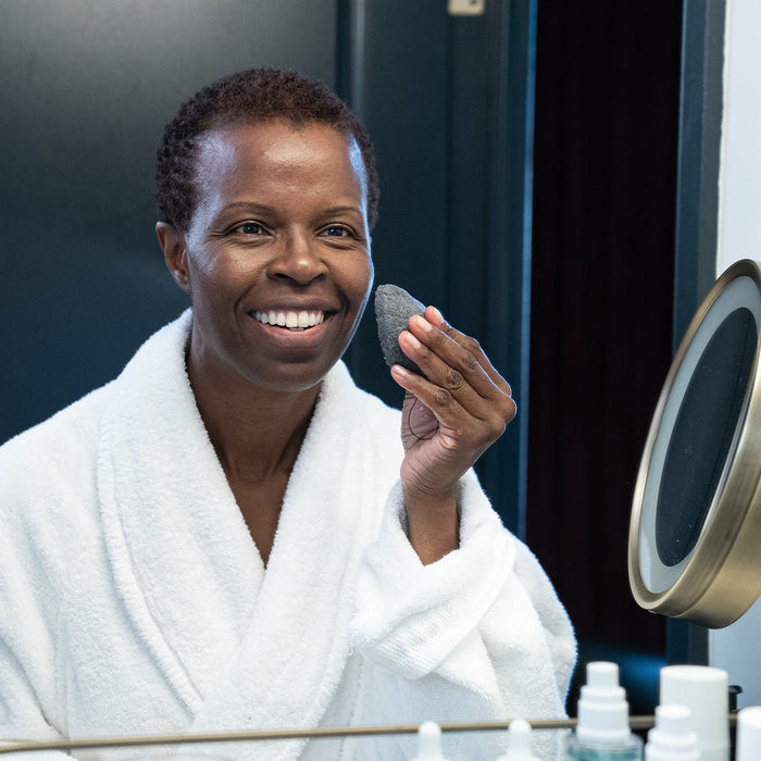 A dark-skinned woman exfoliating her skin with a konjac sponge for sensitive skin in front of a mirror
