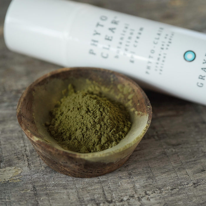 A natural face moisturizer containing chlorella in a white bottle next to a small brown wooden bowl of powdered chlorella sitting on a rustic surface. 