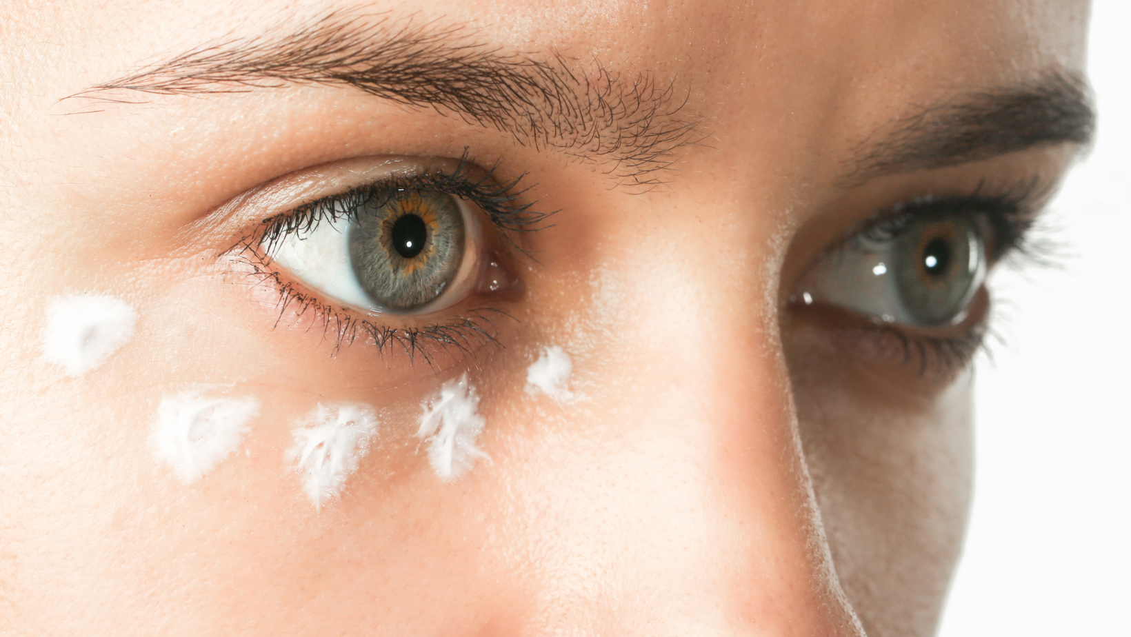 Close up of woman's eyes with dots of white eye cream under one eye.