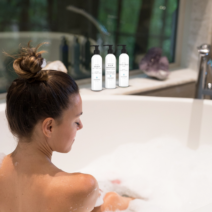 A woman with her hair tied in a messy bun sitting in a white bathtub containing luxurious bubbles with skin and haircare products in the background