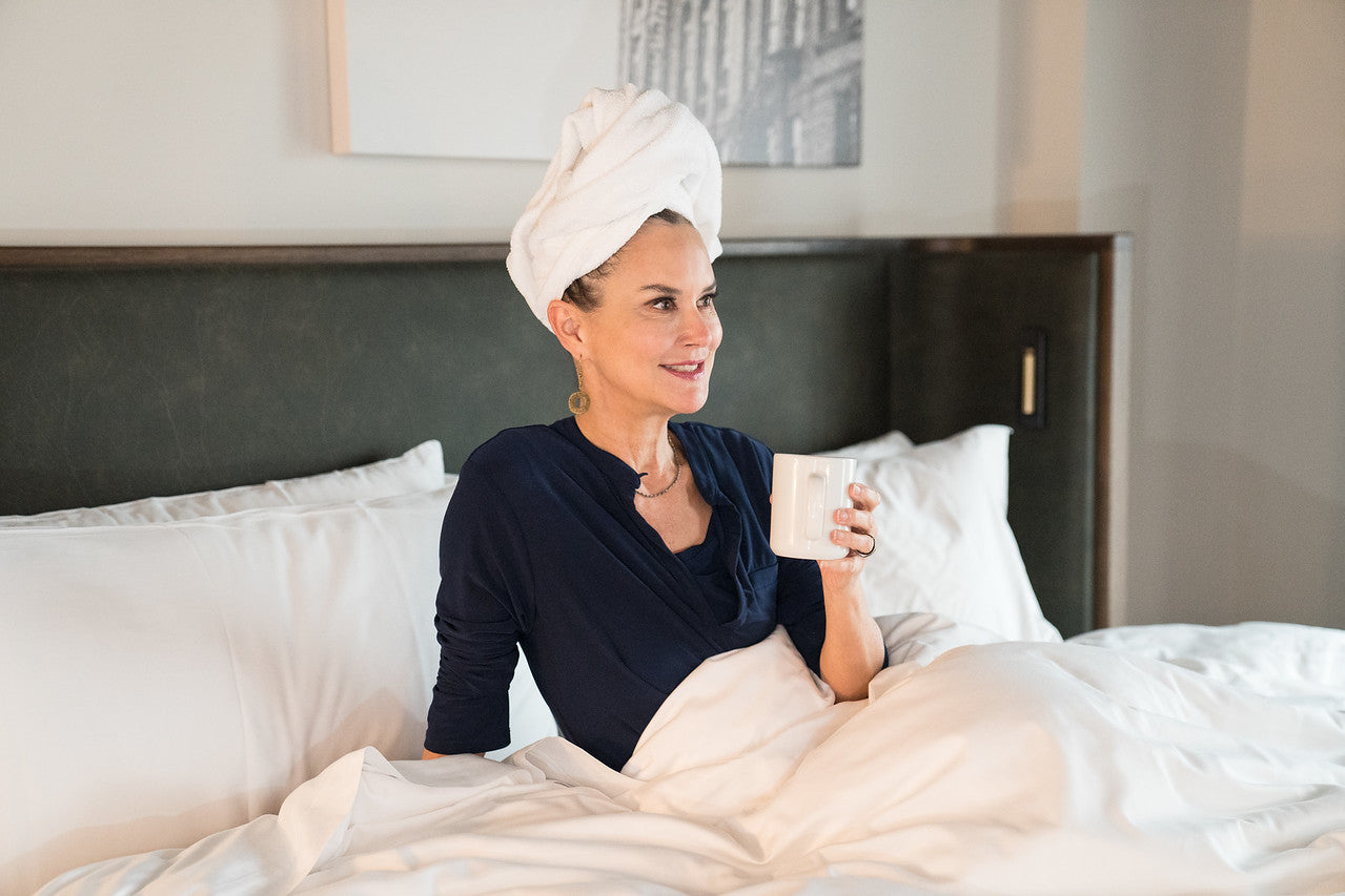 A woman sitting in a hotel bed holding a cup of coffee in her hand with her hair wrapped in a towel