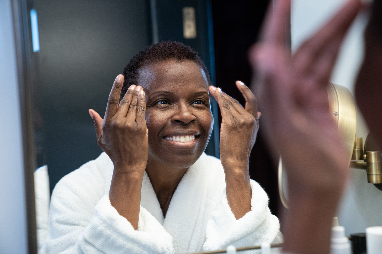 A smiling, dark-skinned woman in a comfortable white robe touching her face with her hands in front of a mirror 