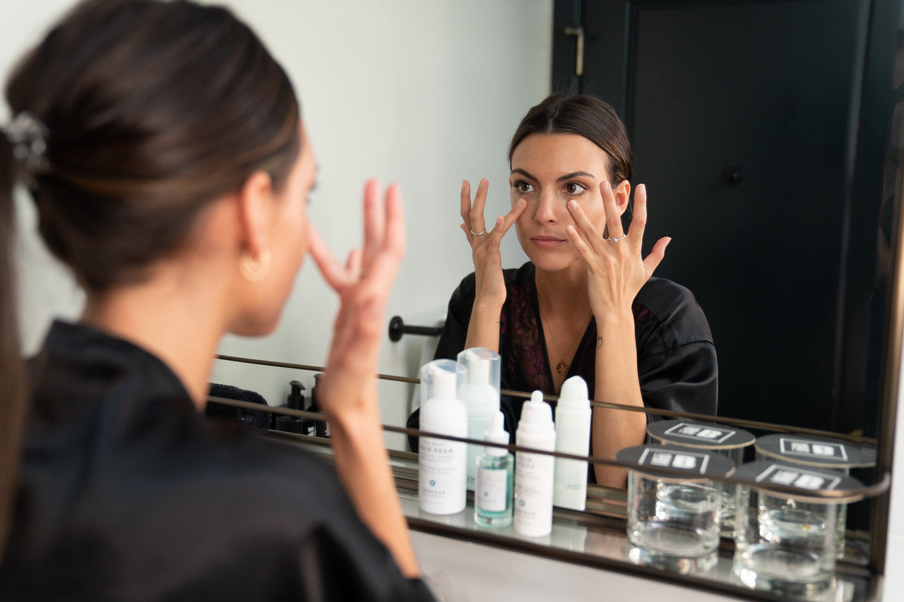 A close-up of woman with her hair tied back in a ponytail applying skincare to her face in front of a mirror using both hands.