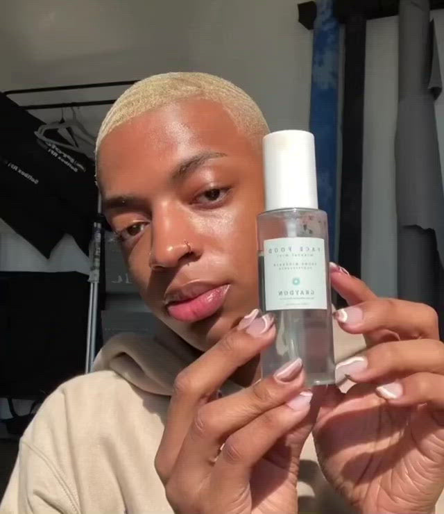 A close-up video of a dark-skinned man misting his face with an aquamarine-coloured vegan hydrating face mist.