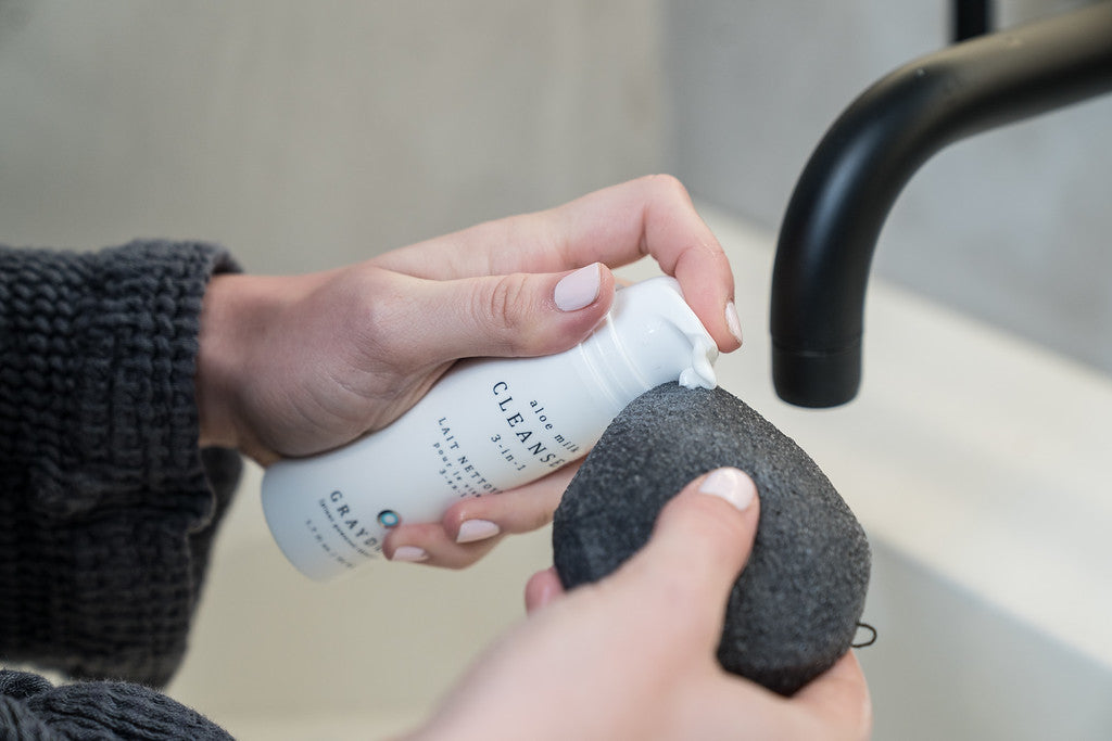 A woman applying a soothing milk cleanser for sensitive skin to a Bamboo Charcoal Konjac Sponge.