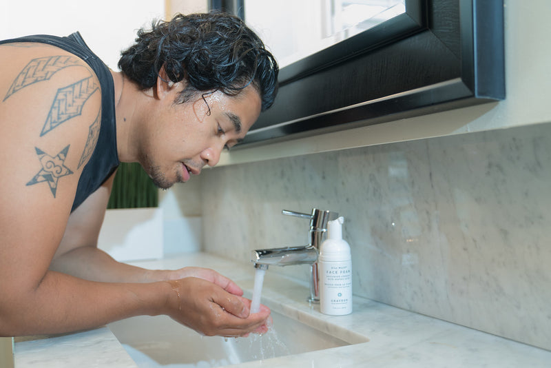 A man rinsing his face with water with a bottle of foaming cleanser for combination skin sitting on his vanity. 