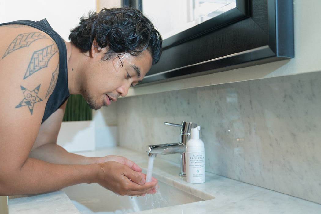 A man rinsing his face with water with a bottle of foaming cleanser for combination skin sitting on his vanity. 