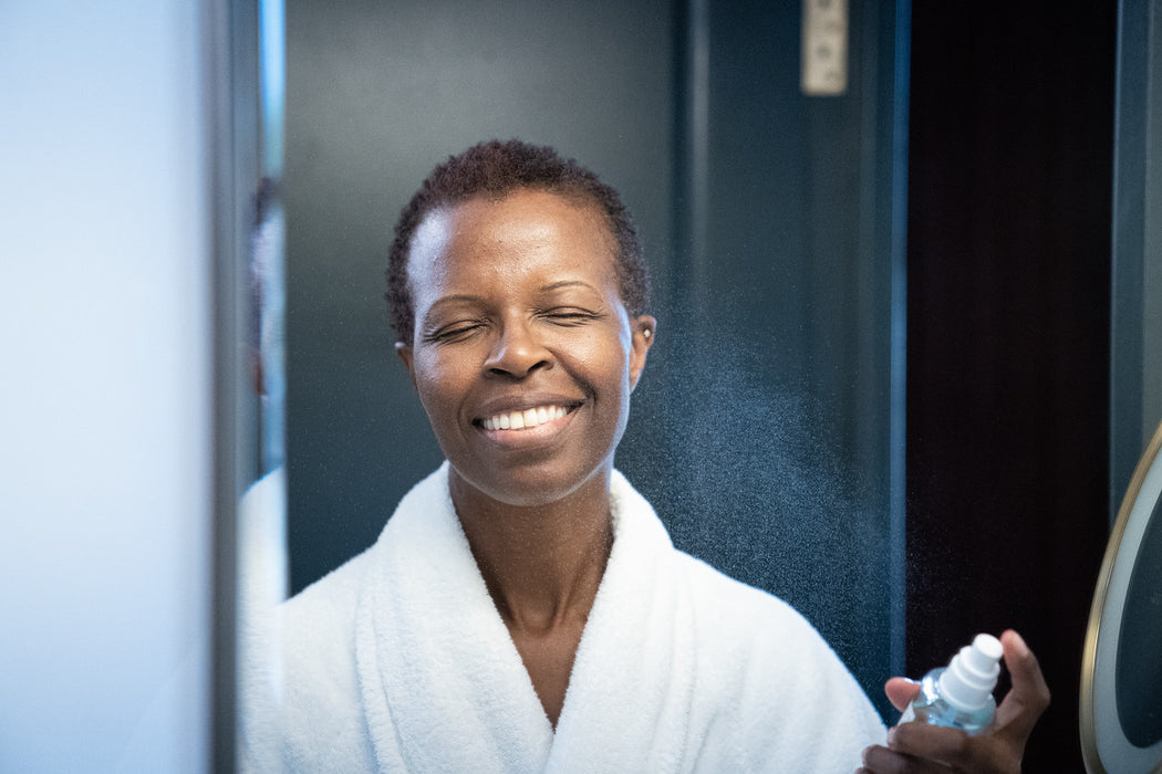  A smiling dark-skinned woman misting her face with an aquamarine-coloured vegan hydrating face mist in front of a mirror. 