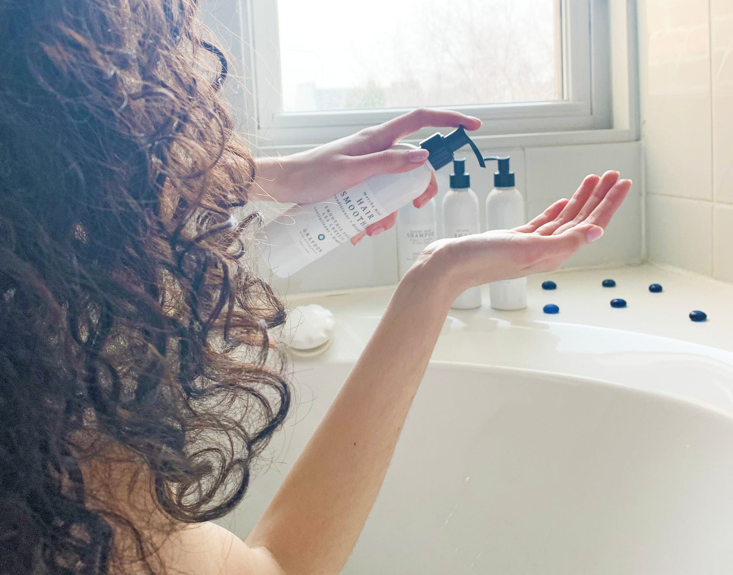A woman with curly hair dispensing a detangling conditioner for sensitive scalp into her hand while sitting in a bathtub. 