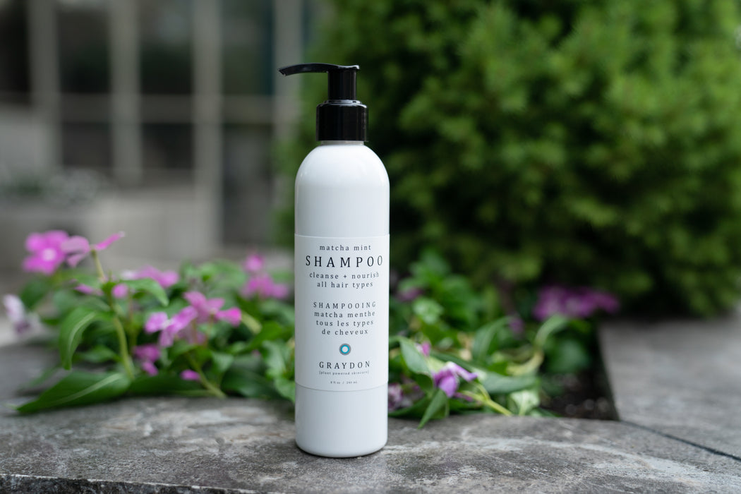 A bottle of moisturizing vegan shampoo in white packaging sitting on a stone ledge with plants in the background. 